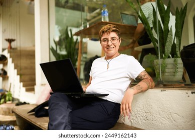 Transgender Gen Z freelancer smiles while working on laptop at eco-friendly coworking space. Inked, digital nomad in casual clothes, emphasizing diversity, inclusivity in modern remote work settings. - Powered by Shutterstock