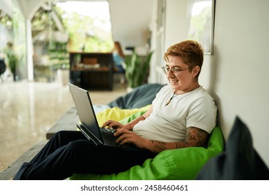 Transgender Gen Z individual smiles while working on laptop seated on green bean bag in airy office. Inclusive, casual pro environment, gender diversity, and modern work culture scene. - Powered by Shutterstock