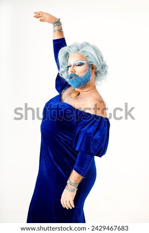 Transgender individual with a dramatic pose, adorned in a luxurious blue velvet dress and striking theatrical makeup, complete with a bold blue beard and stylized eyebrow detailing