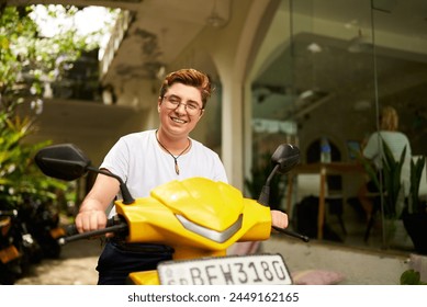 Transgender individual smiles atop yellow scooter outside coworking space, embracing urban eco commute. Joyful moment, modern work-life balance with transport, represents diverse lifestyles. - Powered by Shutterstock