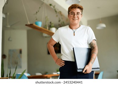 Transgender professional in bright office smiles, holding laptop, inked arms visible. Confident stance inclusive work culture advocate. Nerdy glasses smart casual work attire. Plants modern workspace. - Powered by Shutterstock