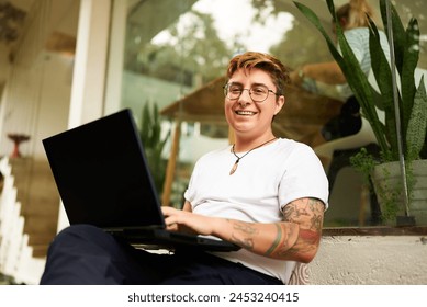 Transgender professional works on laptop, smiles in modern coworking space. Inclusive workplace, creative freelancing, comfy setting. Tattooed, gender diversity, productive remote working. - Powered by Shutterstock