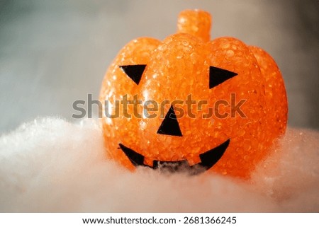 Translucent orange jack-o’-lantern with a textured bubble surface sits on artificial snow. Neutral gray background enhances its warm glow, blending Halloween charm with a hint of winter magic.