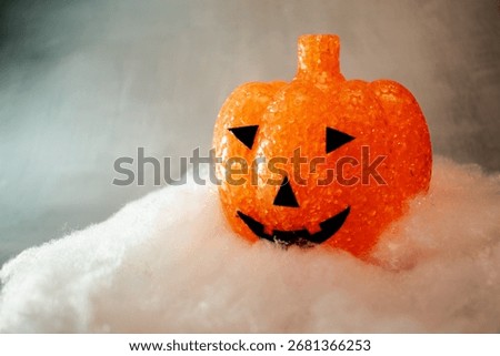 Translucent orange jack-o’-lantern with a textured bubble surface sits on artificial snow. Neutral gray background enhances its warm glow, blending Halloween charm with a hint of winter magic.
