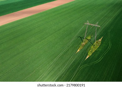 Transmission Tower Supporting Overhead Power Line Through Cultivated Crops Field, Aerial View From Drone Pov