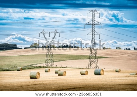 Transmission towers and power lines overlooking a farm field during fall harvest with round hay bales on the Canadian prairies in Rocky View County Alberta Canada.