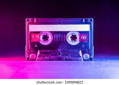 Transparent audio cassette tape lit by pink and blue lamps on a black background. Front, top view.