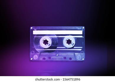 Transparent audio cassette tape lit by pink and blue lamps on a black background. Front, top view.