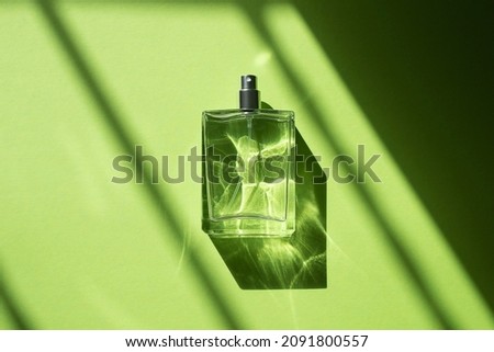 Transparent bottle of perfume with spray on a green background. Natural light and geometric diagonal shadows. Clear glass without lid. Fragrance presentation with daylight. Women's and men's essence.