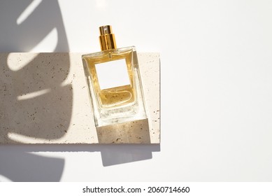 Transparent bottle of perfume with white label on stone plate on a white background. Fragrance presentation with daylight. Trending concept in natural materials with palm leaf shadows.