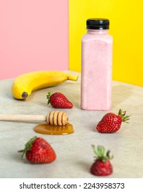 A transparent bottle of strawberry with milk smoothie. The background is yellow and pink. There are strawberries and a banana and honey in the photo, too.