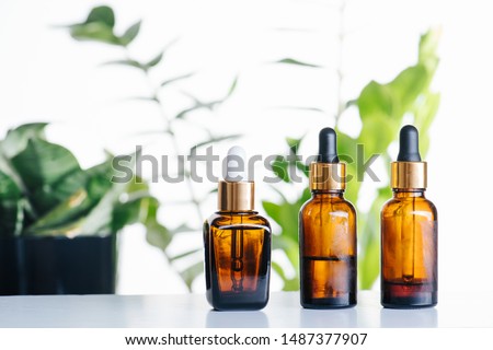Transparent cosmetic amber glass dropper bottles over green plants on white background. Vials with pipette plastic caps for essential oils, perfumes and skincare substances. Three in a row.