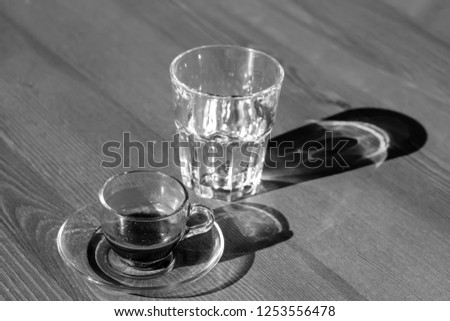 A transparent cup of coffee ground and an empty  glass stand on the table. The morning sun shines on the cup and glass, their shadows on the table. Black and white.