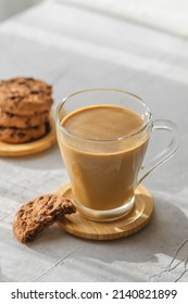 a transparent cup of coffee with milk with chocolate cookies at morning shadows and sun rays through the window on gray table background. Morning coffee concept, close up