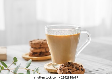 a transparent cup of coffee with milk with chocolate cookies and eucalyptus leaves at morning shadows and sun rays through the window on gray table background. Morning coffee concept, close up