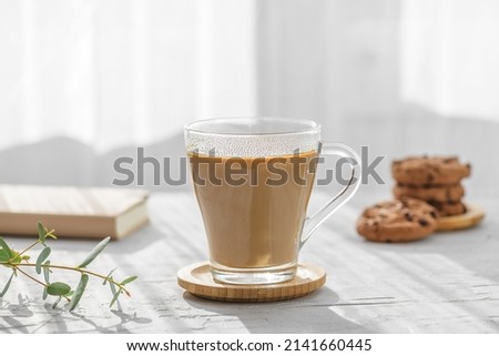 a transparent cup of coffee with milk with chocolate cookies and eucalyptus leaves at morning shadows and sun rays through the window on gray table background. Morning coffee concept, close up