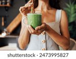 A transparent cup with a green smoothie in the hands of a young woman. A girl in a linen sundress drinks a fresh drink made from herbs and vegetables in the kitchen. Food and drinks are useful.