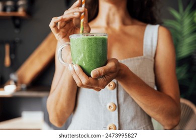 A transparent cup with a green smoothie in the hands of a young woman. A girl in a linen sundress drinks a fresh drink made from herbs and vegetables in the kitchen. Food and drinks are useful. - Powered by Shutterstock