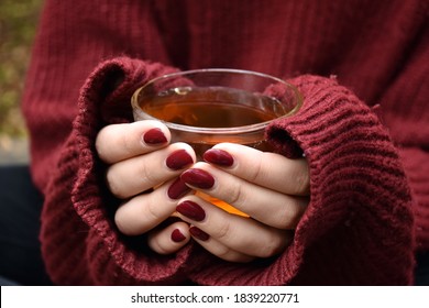 A transparent cup with tea in the hands of a young girl in a brown sweater.