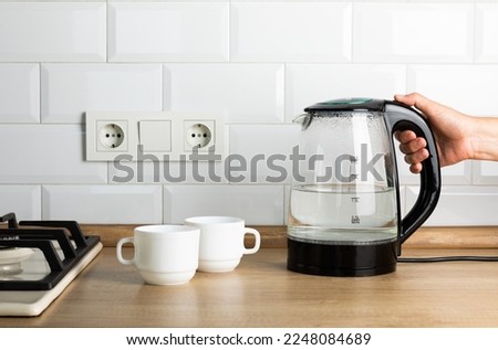 A transparent electric kettle with boiling water in a woman's hand and cups for tea on the table in the kitchen. The woman is making tea.