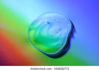 Transparent Gel Drop On Neon Rainbow Light Overlay Background. Top View. Virus Protection Or Cosmetics Concept. Serum Texture Close Up.