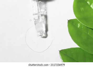 Transparent Gel Pouring Onto A White Surface From A Pipette, Top View. Essence Of Cosmetic Product With Green Leaves Of The Plant, Body Care.