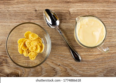 Transparent Glass Bowl With Corn Flakes, Spoon, Pitcher With Fruit Yogurt On Brown Wooden Table. Top View