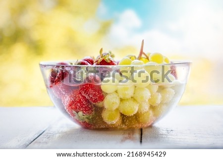 Transparent glass bowl filled with freshly picked strawberries and grapes, washed in water. Blurry bright sky and trees in the background. Summer organic farm fruits.