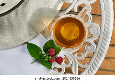 Transparent Glass Cup Of Herbal Tea And Sun Hat On A Garden Table  In The Garden. White Book With Red Raspberries. White Hat On A White Garden Table. Shallow Depth Of Field. Top View