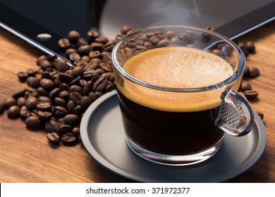 A Transparent Glass Cup Of Hot Steamy Espresso On A Saucer Standing On A Wooden Table Top, Surrounded By Coffee Beans And An IPad In The Background