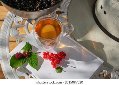 Transparent Glass Cup Of  Tea And Sun Hat On A Garden Table In The Garden. White Book With Raspberries. White Hat On A White Garden Table. Shallow Depth Of Field. Top View