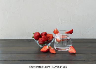 Transparent glass of water with slice of strawberries, cutted starwberries and berries in little plate and on dark wooden table, copy space. Refreshing vitamin detox concept