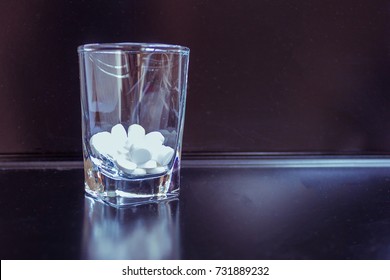 In A Transparent Glass Are White Tablets. A Glass Is Reflected In The Black, Care-giving Surface Of The Table. Dark Background.