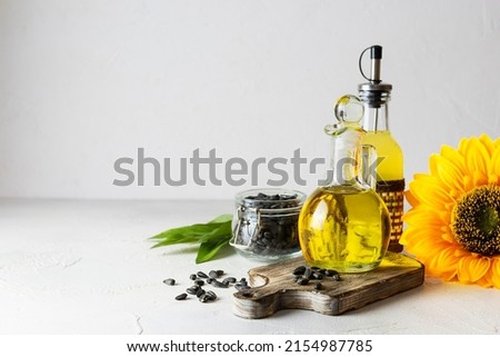 Transparent jug with sunflower oil on a dark wooden plank. A bottle of butter and seeds in a glass jar in the background. Light background, space for text