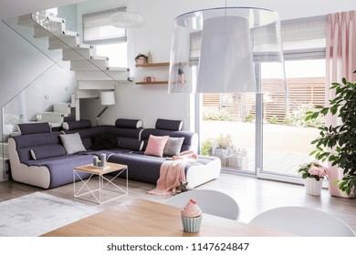 Transparent Light Shade Above A Wooden Dining Table In A White Living Room Interior With A Modern Corner Sofa And Stairs With Glass Barricade