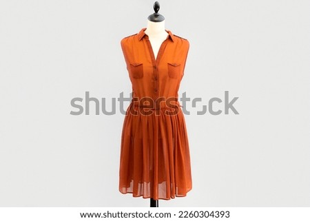 A transparent orange shirtdress dress with buttons hangs on a headless mannequin. isolated by a light gray white background.