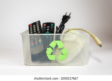 Transparent Plastic Box With The Recycling Symbol Containing Electronics Such As Tv Remotes, Cables Or An Old Computer Mouse On A White Background