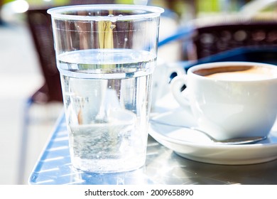 Transparent plastic glass with water with cup of cappuccino and caffe tables in background blurred. Traditional italian style of serving coffee. Still drinking water vs caffeine. Healthy dieting. 
