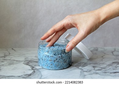 Transparent Plastic Open Jar With Homemade Lavender Sugar Scrub Dyed Blue. Beautiful Female Hand With Graceful Fingers Is Lowered, Touches The Scrub In Jar With One Finger. Image With Selective Focus