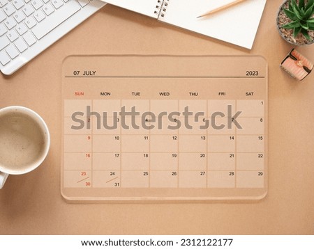 Transparent tablet computer with an app of the calendar for July 2023 year and a cup of coffee, keyboard, notebook, and pencil on brown background. Concept schedule planning. Top view, flat lay.