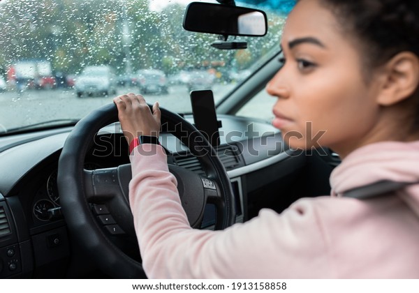 Transport, journey and travel in city with gadget.
Serious young african american lady with seat belt, driving car and
looking to side, next to smartphone with empty screen, side view,
copy space