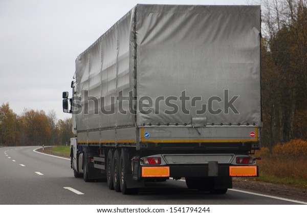Transport Logistics,\
international commercial freight by road, gray awning semi truck\
drive next to the two-lane asphalted country road in the autumn\
day, close up side rear\
view
