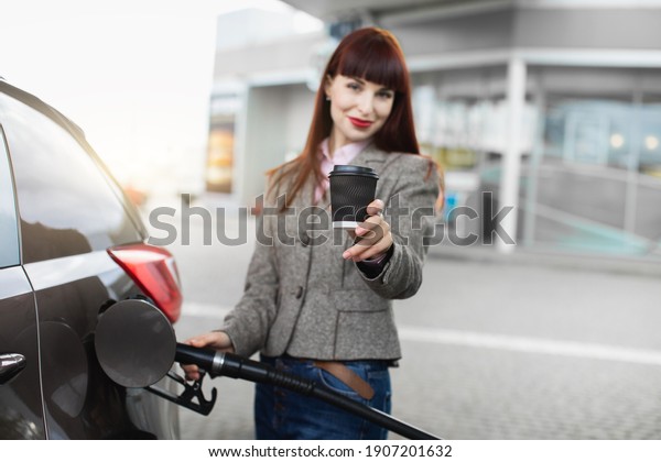 Transportation, technology, energy concept. Blurred\
portrait of young pretty businesslady, refueling her car at gas\
station, and demonstrating to camera paper cup of coffee to go.\
Focus on the\
cup