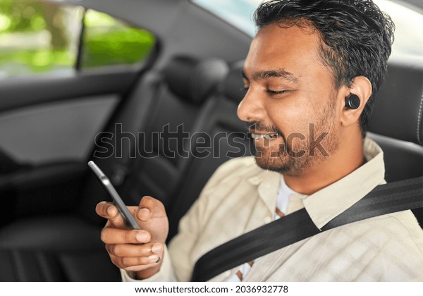transportation, technology and people concept - happy\
smiling indian male passenger with wireless earphones using\
smartphone on back seat of taxi\
car