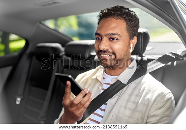 transportation, technology and people concept - happy\
smiling male passenger with wireless earphones using smartphone on\
back seat of taxi\
car