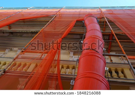 Trash chute or debris chute in red color on a facade of a historic building in reconstruction for rubbish removal. The house is covered with red protective net. The photo is taken at low angle.