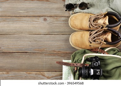 Travel Accessories Set On Wooden Background: Old Hiking Leather Boots, Pants, Backpack, Map, Vintage Film Camera And Sunglasses. Top View Point.