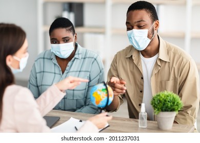 Travel Agent And Black Family Couple Choosing Destination For Vacation Pointing At World Globe Sitting In Modern Office Indoors, Wearing Protective Face Masks. Tour Agency Service. Selective Focus