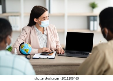 Travel Agent Lady Showing Laptop With Empty Screen To Family Couple Offering Vacation Tour Sitting At Desk In Modern Office, Wearing Face Mask. Mockup For Touristic Website Concept