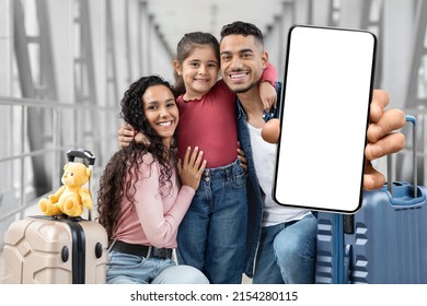 Travel App. Cheerful Middle Eastern Family At Airport Demonstrating Blank Smartphone At Camera While Waiting For Their Flight At Terminal, Parents And Child Recommending New Application, Mockup